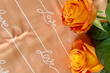 Load image into Gallery viewer, closeup shot of Love script on Peach Fuzz background with orange roses on right side
