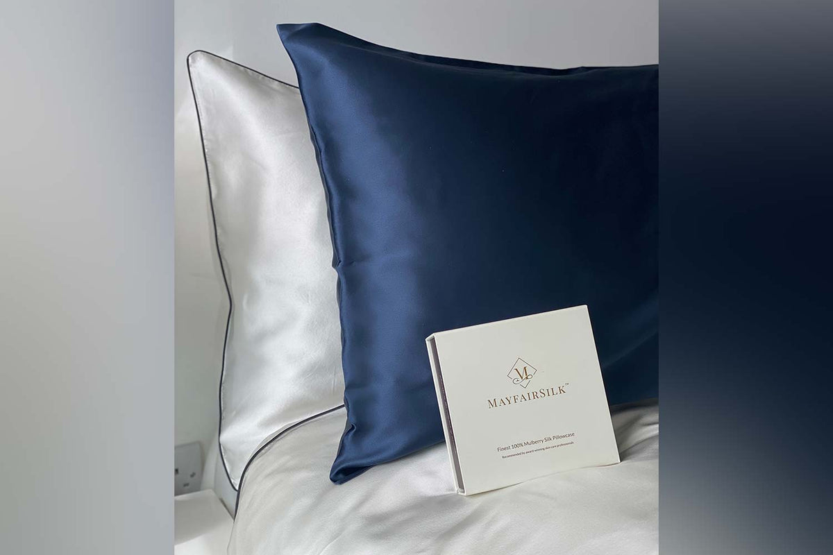 Midnight Blue Silk pillowcase sitting upright in front of a white silk pillowcase with charcoal piping on the border, with a gift box sitting in the foreground. 